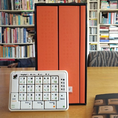 the book "shift happens" in its box on a table in front of bookshelves, a small frogpad keyboard in front of it