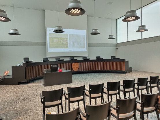 Gerichtssaal, mit Schweizerkreuz in der Mitte der Richter*innenbank. Im Saal sind Stühle in Reihe gestellt.