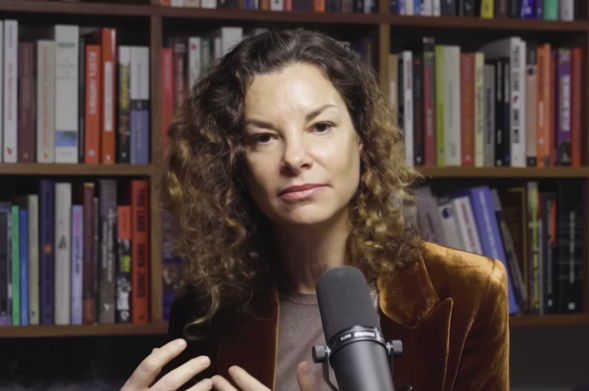 Gaia Vince, long brown curly hair,  interviewed in front of a bookshelf which takes the complete background