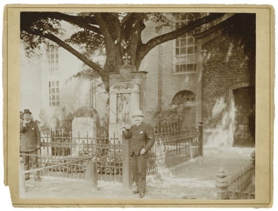 Zwei Männer stehen vor einem Grab, das von einem schmiedeeisernen Zaun umgeben ist. Ein Mann ist in Anzug und Hut und zeigt auf das Grab, während der andere Mann im Hintergrund steht, teilweise von Schatten verdeckt. Es gibt einen großen Baum in der Bildmitte.
