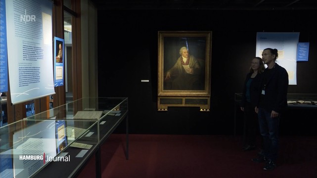 Ausstellungsraum mit Vitrinen, Informationstafeln, einem großen gerahmten Klopstock-Porträt und zwei Personen, die beobachten. Logo 