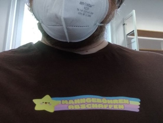 Selfie einer Person. Oberkörper in einem braunen T-Shirt und mit Maske verdecktem Mund sind zu sehen. Auf dem T-Shirt ist ein Bügelbild des Mahngebühren-Abschaffen-Stickers (Stern mit buntem Schweif auf dem der Schriftzug steht).