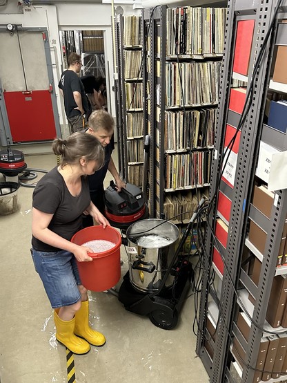 Leute, die einen überfluteten Archivraum reinigen. Eine Person hält einen roten Eimer, und eine andere betreibt ein nasses Vakuum. Die Regale sind mit Vinyl-Schallplatten gefüllt.