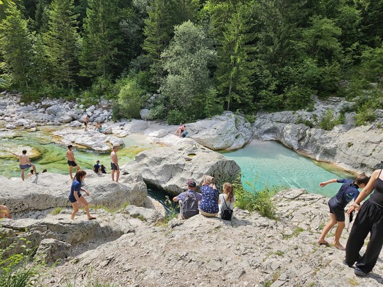 Menschen stehen an Felsen, von denen man in einen türkisfarbenen Fluss springen kann. 