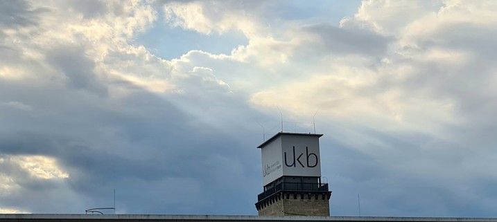 Wolken mit Sonnenstrahlen über einem Turm, an dem ukb für Universitätsklinikum Bonn steht.