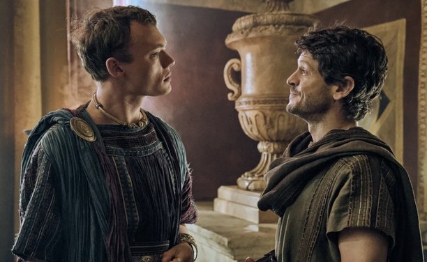 Two men in ancient Roman attire are conversing indoors with an ornate urn in the background.
