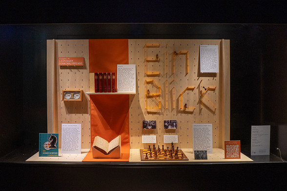 Eine Ausstellungsvitrine, darin eine Holzlochplatte mit dem Ausstellungsobjekten: Buch, Schachuhr, Schachbrett mit Figuren, Fotos