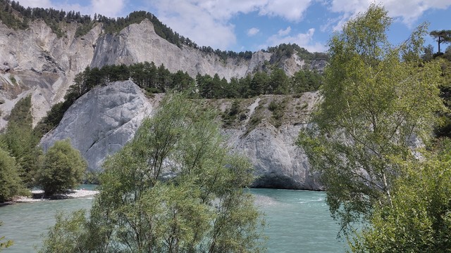 Blick auf eine Fluss, dahinter ein Berg. Der Fluss ist türkis-blau, der Berg besteht aus steilen Felsen, darauf einige Bäume.