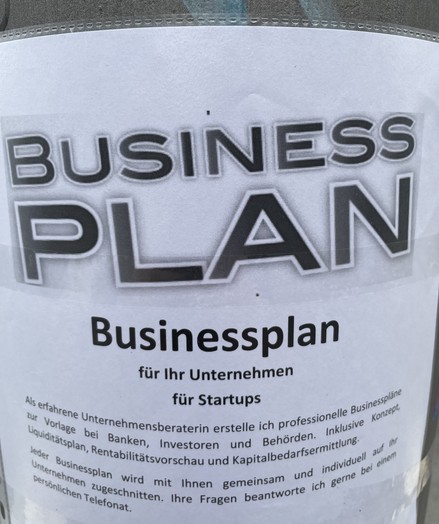 Foto einer an einer Laterne angebrachten Werbung. Oben ist eine WordArt-basierte Überschrift „Business Plan“ zu sehen, unten folgt ein Text, der die Professionalität der Unternehmensberaterin bewirbt.