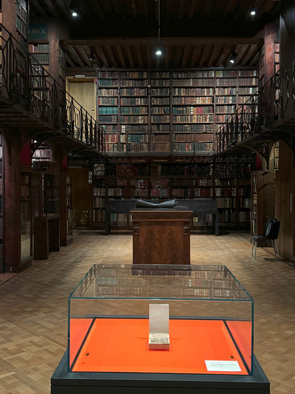 Blick auf die Vitrine in deren Zentrum das Antwerpener Liederbuch aufgeschlagen liegt. Die Vitrine befindet sich in einem weitläufigen Bibliotheksraum der mit dunklen Holzregalen befüllt ist.