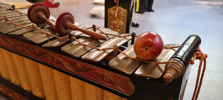 Gender eines javanischen Gamelan. Auf einem geschnitzten Ständer sind mit einer Schnur Metallplatten befestigt. Unter den Platten sind Bambusrohre ausrecht aufgestellt, die die Resonanz verstärken. Obendrauf liegen die Schläger, die zu diesem Instrument gehören und ein Apfel.