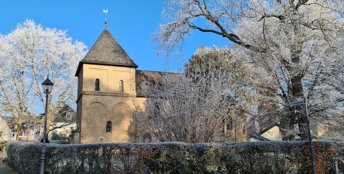 Eine romanische Kapelle in Köln, genannt Krieler Dömchen, umgeben von einer Hecke und Bäumen, die von Raureif bedeckt sind. Strahlend blauer Himmel und Sonnenschein.