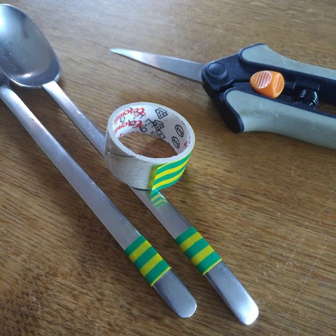 foto zweier edelstahl-löffel mit einer markierung mit grüngelb gestreiftem klebeband am oberen teil des griffs, daneben eine schere und die klebeband-rolle, die fast leer ist