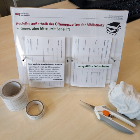 foto des A4-schildhalters mit einem infobogen zur bibliotheksausleihe und den beiden A6-prospekthaltern "draufgeklebt" mit leihscheinen
