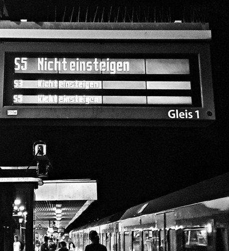 Black and white photo of a train platform display board. Three lines repeatedly show 'Do not board' for S-Bahn lines. 'Platform 1' at the bottom. Silhouettes of a train and passengers in the foreground. 