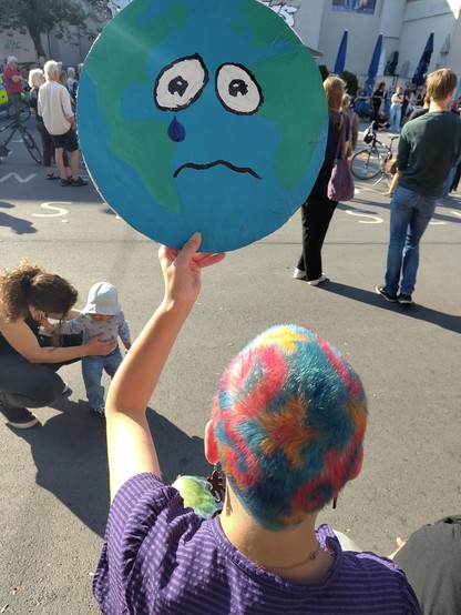 Eine Person mit bunten, kurzen Haaren hält ein Pappschild hoch, auf dem eine traurige Erde abgebildet ist. 