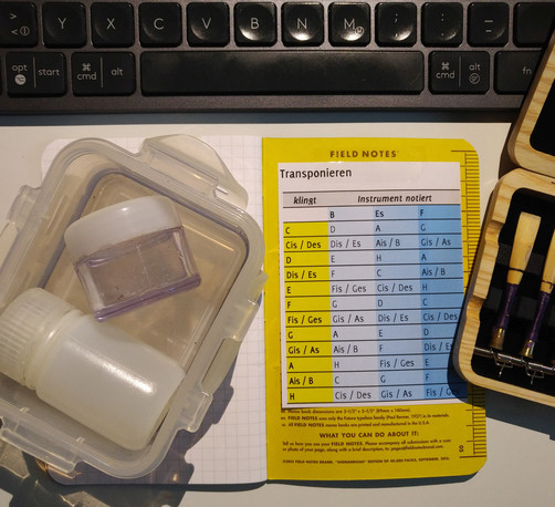 foto einer kleinen eckigen kunststoffdose, in der zwei kleine runde kunststoff-dosen mit wasser drin liegen, eine transponiertabelle, die angibt, welche in C klingenden töne welche töne auf instrumenten sind, die in B, Es oder F gestimmt sind, und rechts am rand sieht man noch ein etui für englischhorn-rohre.