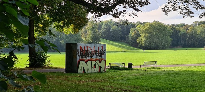 Bretterbude im Park für mobile Toiletten. Rückseite mit den Buchstaben NDH besprüht, einmal ganz einfach und mit schwarzen Klecksen übermalt und ein zweites Mal in kunstvoller Ausführung in Gelb mit schwarzen und roten Schatten.