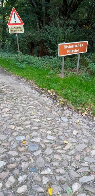 Straße in Brunsbrock mit historischem Pflaster und Hinweisschild darauf, zusätzlich Verkehrszeichen: Achtung, schlechte Wegstrecke.