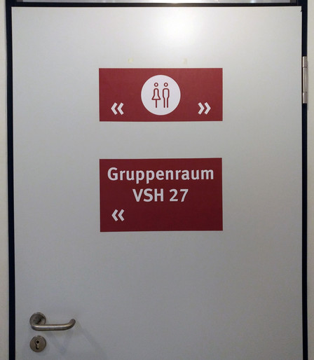 foto einer hellgrauen tür, darauf zwei rote hinweisschilder auf toiletten zu beiden seiten der tür und zu einem gruppenraum namens 