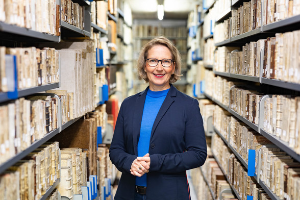 Porträt von Prof. Dr. Caroline Emmelius. Sie hat kinnlange blonde Haare und eine Brille. Sie trägt einen dunkelblauen Blazer und einen blauen Pullover darunter. Sie steht inmitten zweier Magazinregale. In diesen sind die Buchrücken von Handschriften zu sehen. Foto: © Christian Klenk