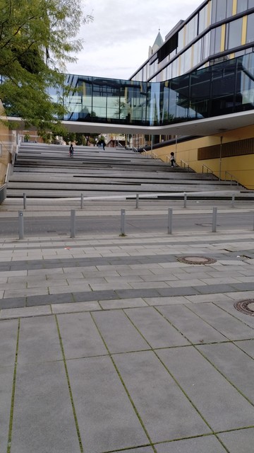 Eine lange, sehr breite und recht flache, mit Betonsteinen gepflasterte Treppe im öffentlichen Raum. Sie hat mehrere Absätze und diagonal durch die Treppenstufen läuft im Zickzack eine Rampe.