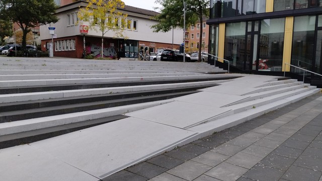 Ein Foto von derselben Treppe, auf dem man ein paar Treppenstufen und die dadurch von links unten nach rechts oben verlaufende Rampe aus geringerer Entfernung sieht