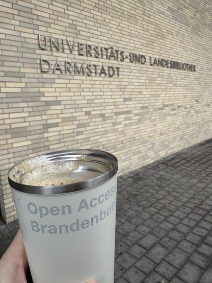 Das Gebäude der ULB Darmstadt, davor meine Hand, die einen Thermobecher mit der Aufschrift “Open Access Brandenburg” hält