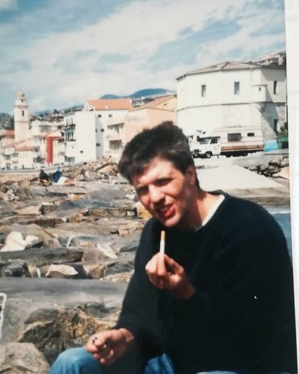 Junger Mann mit einer Zigarette in der Hand. Im Hintergrund sind Häuser in einer Stadt in Ligurien in Italien.
