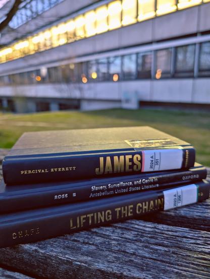 A stack of 3 black books with golden writing on the spine lie on a wooden bench outside of SUB Göttingen, with the dusky sky and an illuminated building in the background. The books are: Pervial Everett, "James"; Kelly Ross, "Slavery, surveillance, and genre in Antebellum United States literature" and William H. Chafe, "Lifting the Chains"