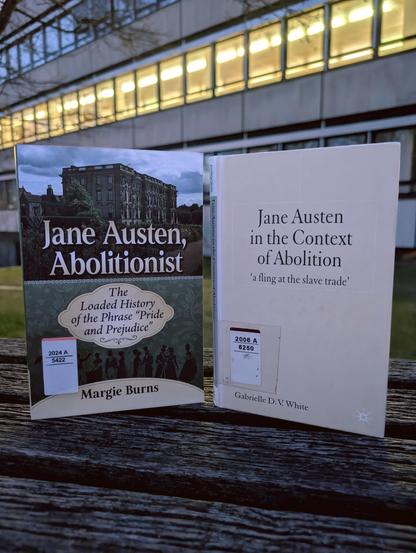 2 books are positioned on a wooden bench outside of SUB Göttingen, with the dusky sky and an illuminated building in the background. The books are: Margie Burns, "Jane Austen, Abolitionist" 
& Gabrielle D V White "Jane Austen in the Context of Abolition"