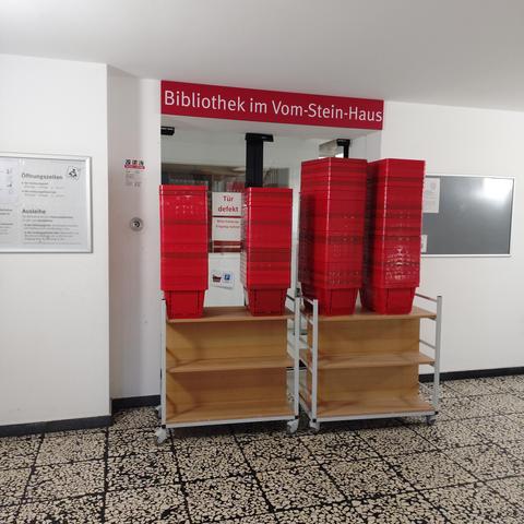 foto einer schiebetür zu einer bibliothek, die, da sie defekt ist, von einem rollwagen mit körben für die bibliotheksbenutzer:innen "abgeschirmt" ist