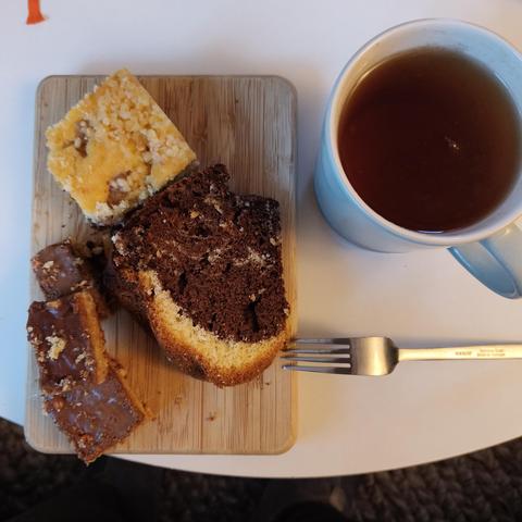 foto einiger kuchenstücke auf einem holzbrettchen auf einem weißen tisch, daneben ein becher tee.