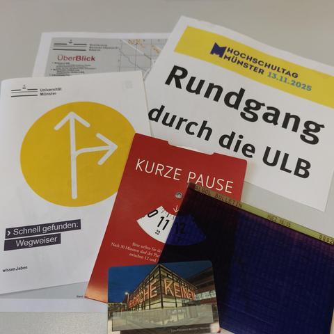 foto einer sammlung von materialien für eine bibliotheksführung: hinweissschild für den treffpunkt, uni-lageplan, wegweiser-handout, "parkscheibe" für arbeitsplätze, ein microfiche-bogen, ein muster-bibliotheksausweis