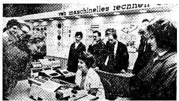 Das Bild ist ein Schwarzweißfoto von fünf Personen, die um einen Tisch versammelt stehen. Der Tisch ist mit verschiedenen Gegenständen bedeckt, darunter Papiere, kleine Schachteln und andere kleine Objekte. Zwei Personen stehen an einer Seite des Tisches und zwei stehen an der anderen. Eine fünfte Person steht vor dem Tisch. Im Hintergrund ist eine Wand zu sehen. Auf der Wand steht "veb maschnelles rechnen".