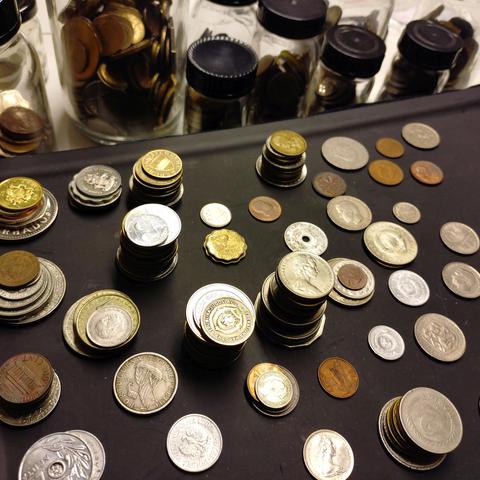 foto eines schwarzen tablets mit stapeln von kursmünzen aus unterschiedlichen ländern, im hintergrund gläser mit weiteren münzen