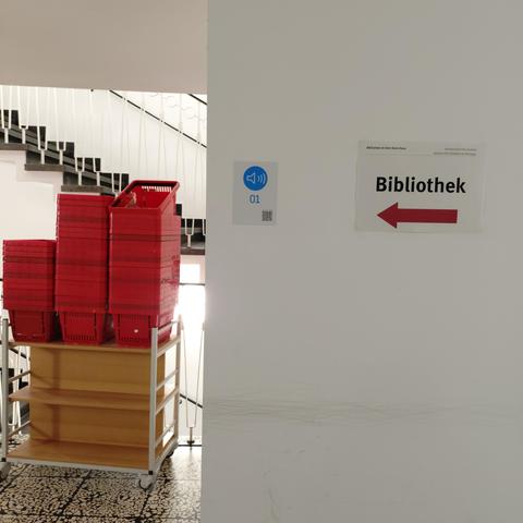 foto eines ausschnitts aus einem treppenhaus mit einem schild, das mit einem pfeil nach links zu einer bibliothek weist, und einem stapel mit körben für die benutzer:innen