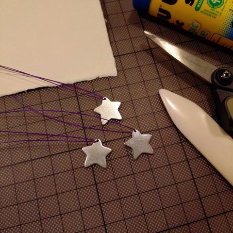 foto von drei silberfarbenen metallenen kleinen sternen mit lila befestigungsband auf einer bastelunterlage; um sie herum liegen papier, ein falzbein, eine schere und ein klebestift