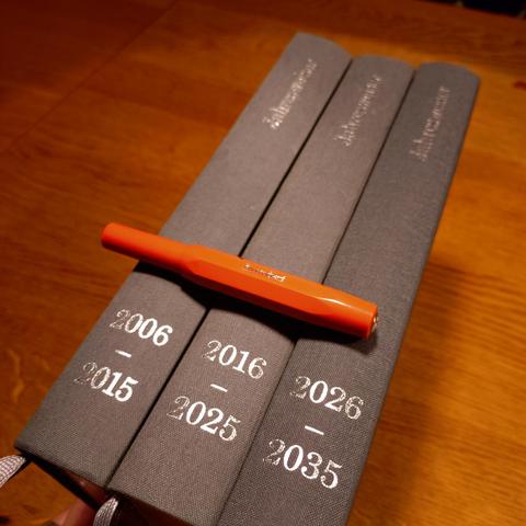 foto der rücken von drei zehn-jahres-kalender-büchern in grauem leineneinband mit silbener prägung der jahreszahlen; auf ihnen liegt ein orangener kaweco-sport-füller