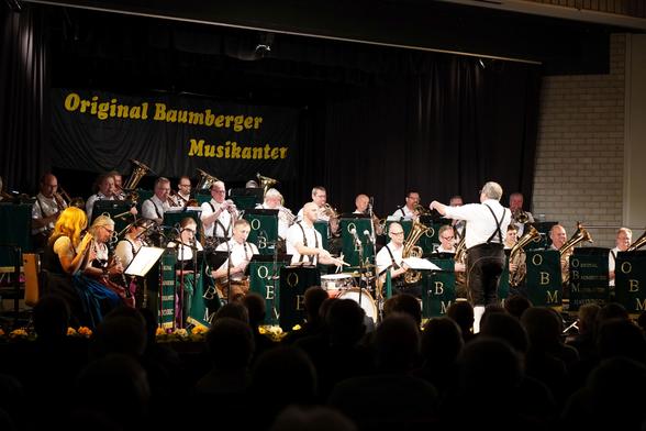 foto eines orchester – den baumberger musikanten aus havixbeck im münsterland – auf einer bühne, im hintergrund ein banner mit dem namen der gruppe
