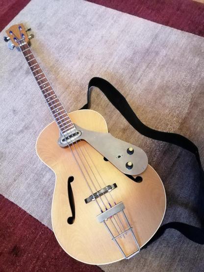 Ein Short Scale Bass, der an eine Jazz-Archtopgitarre erinnert: Hohlkorpus mit F-Löchern naturfarben, schlanker Basshals, schwebend am Halsende montierter Pickup mit Metall-Pickguard. Am Bass ist ein einfacher Gurt montiert, das Instrument liegt auf einem Wollteppich.