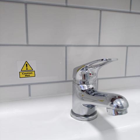 foto eines waschbeckens in einem weiß gefliesten badezimmer; schräg hinter dem wasserhahn an der wand klebt ein aufkleber mit einem gelben dreieck mit einem ausrufezeichen und darunter der aufschrift "caution hot"