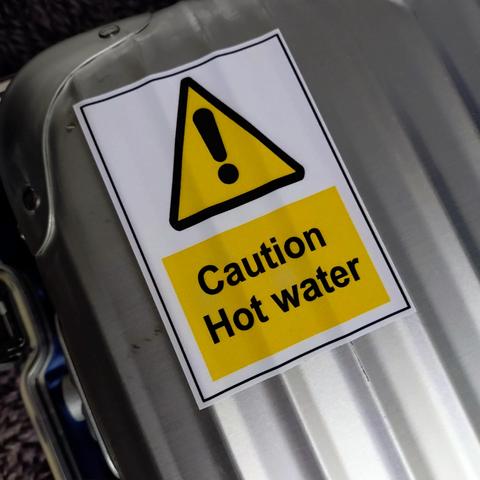 foto eines aufklebers mit einem gelben dreieck mit einem ausrufezeichen und darunter der aufschrift "caution hot water" auf einem aluminiumkoffer