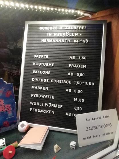 Schaufenstersuslage des Zaubereifachgeschäfts "Zauberkönig" in Berlin-Neukölln mit einer Tafel der geführten Produkte: Bärte, Kostüme, Ballons, diverse Scheisse.