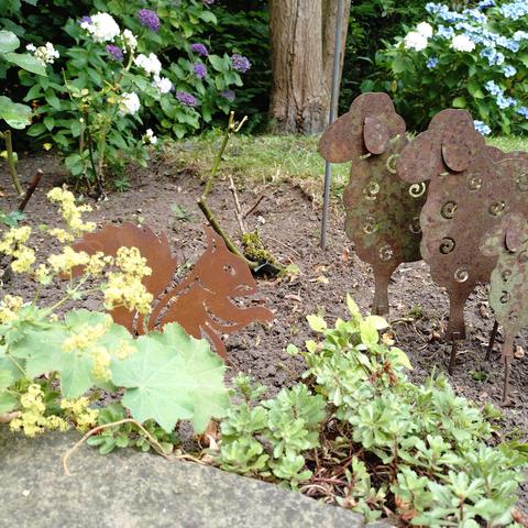 fotos eines blumenbeetes, in dem eine flache eichhörnchen-figur setzt, beobachtet von drei schaf-figuren von rechts