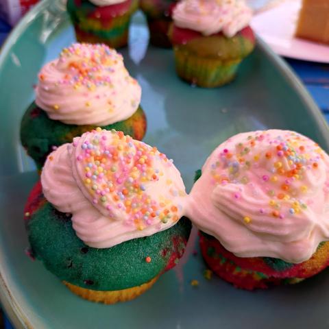 foto eines tellers mit einigen muffins in regenbogenfarbenem teig mit sahnehaube und bunten zuckerstreuseln