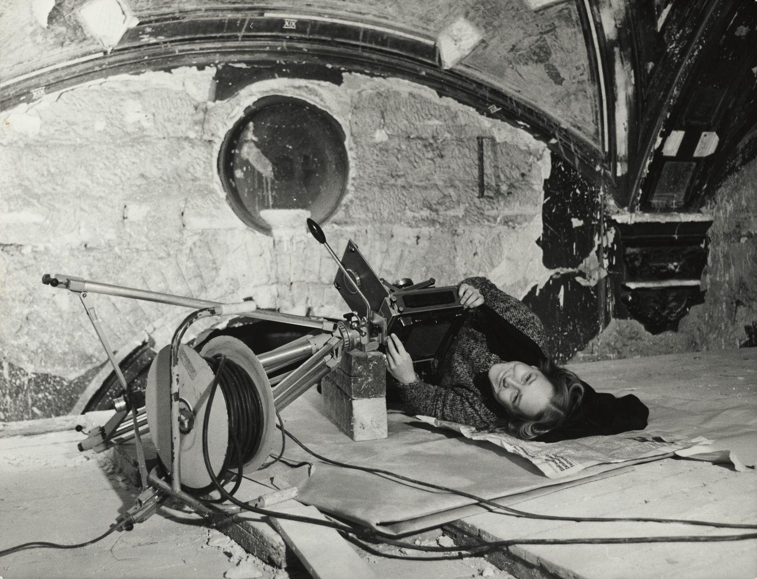 Schwarz-Weiß-Foto von Regine Richter, die während der Rekonstruktion der Semperoper flach auf einer Zeitungsunterlage auf dem Boden liegt, um eine tief positionierte Spezialkamera zu bedienen.