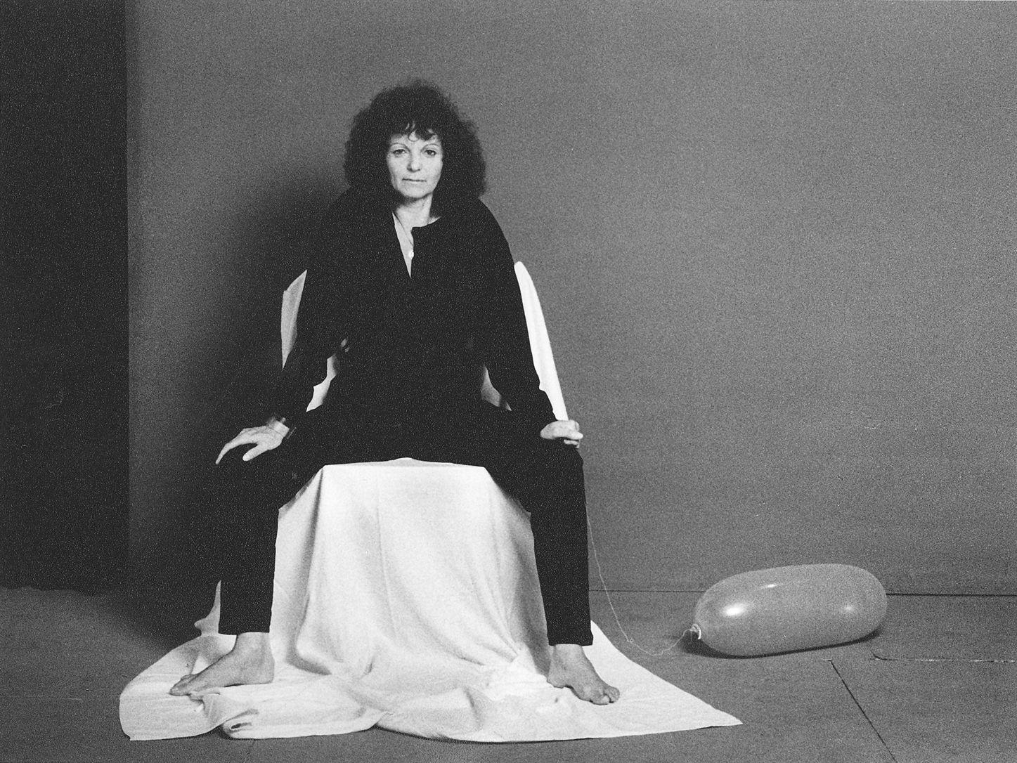 Schwarz-Weiß-Selbstporträt von Evelyn Krull, die barfuß und in dunkler Kleidung auf einem weiß verhüllten Stuhl sitzt, während rechts auf dem Boden ein länglicher Luftballon liegt.